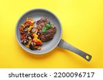 Tasty fried steak with vegetables in pan on yellow background, top view
