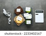 Calculator and food products with calorific value tags on dark grey table, flat lay. Weight loss concept