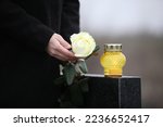 Woman holding white rose near black granite tombstone with candle outdoors, closeup. Funeral ceremony