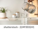 Woman pouring water from jug into glass at white table in kitchen, closeup. Space for text