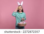 Happy woman in bunny ears headband holding wicker basket of painted Easter eggs on pink background