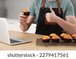 Man holding muffin near laptop and showing thumb up at table in kitchen, closeup. Time for hobby