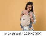 Small photo of Mother holding her child in sling (baby carrier) on beige background. Space for text