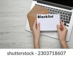 Small photo of Woman holding letter with word Blacklist near laptop at white wooden table, top view. Space for text
