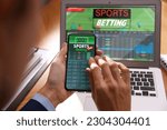 Man betting on sports using smartphone and laptop at table, closeup. Bookmaker websites on displays