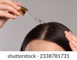Small photo of Woman applying essential oil onto hair roots on light grey background, closeup