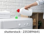 Woman pouring fabric softener from bottle into cap on washing machine indoors, closeup