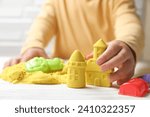 Small photo of Little child playing with yellow kinetic sand at white wooden table, closeup