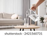 Woman pouring fresh water from jug into glass at wooden table indoors, closeup. Space for text