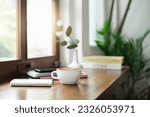 Close-up view, white coffee cup on wooden counter and smart phone, note book put on digital tablet by window, coffee break and relax after work in cafe with a reading book in the background