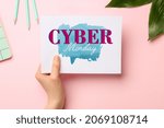 Female hand holding sheet of paper with text CYBER MONDAY on pink background