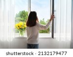 Pretty young Asian woman opening window at home on sunny day, back view