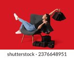 Young Asian woman with gift boxes and shopping bags sitting on chair against red background. Black Friday sale