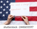 Small photo of Woman writing text WE NEED A CHANGE - IMPEACHMENT NOW on picket poster against USA flag