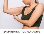 Small photo of Cropped shot of a young caucasian blonde woman grabbing skin on her upper arm with excess fat isolated on a gray background. Pinching the loose and saggy muscles. Taking care of the body