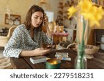Beautiful young woman is guessing on cards with tarot, runes on wooden table and uses an online app in phone to interpret divination, home interior,candles and dry herbs..