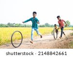 Small photo of happy cheerful Indian village kids playing with tyre wheel rolling near paddy field rural street - concept of entertainment, holidays and leisure activities.