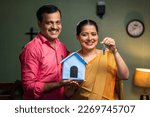 Happy smiling couple showing new house keys by toy home while looking camera - concept of new home purchasing, home loan and investment.