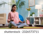 Indian peaceful pregnant woman in lotus pose doing meditation on yoga mat home - concept of mindfulness, relaxation and Self-care during pregnancy
