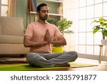 Small photo of Indian young man doing namaste posture or yoga with closed eyes while sitting at home - concept of healthy lifestyle, fitness and self caring