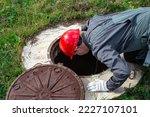 Small photo of A male plumber opened the hatch of a water well and looks inside. Inspection of water pipes and meters.