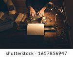 Typing hands of a writer looking for inspiration to start a new novel on his vintage typewriter in a retro style studio: a wooden desk lit by a lamp, an old book and a pair of glasses.