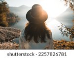 Back view of fashionable young woman with long hair, stylish denim jacket and sleek black hat looking at view against lake reflections and sun beam. Calm water, serene atmosphere, breathtaking scenery