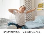 Small photo of Cute little kid boy wake up in the morning in his bed, stretching hand rise up to the air while sitting in sunny bedroom with big window on background.