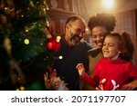 Small photo of Beautiful young mixed race family enjoying their holiday time together, decorating Christmas tree, arranging the christmas lights and having fun