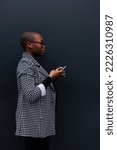 Small photo of An African American woman is standing outdoors in front of a black background wall holding a smartphone in her hands and looking froward.