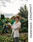 Small photo of Young Caucasian woman holding freshly dug up beet in her fathers garden and showing it to the camera while her father works in the background