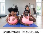Young short curly black hair man and his wife relax on yoga ball while both daughter are on their back. Happy family enjoy holiday together in fitness center.
