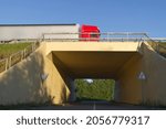 Large transport or haulage lorry passing overhead on the bridge over an empty underpass road with leafy green trees against a blue sky