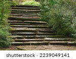 Steep stone staircase with uneven steps on the slope in the landscape design of the park. Fallen leaves are lying on the steps.                       