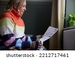 Small photo of Woman In Gloves With Hot Drink And Bill Trying To Keep Warm By Radiator During Cost Of Living Energy Crisis