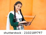 Portrait of Happy young indian school girl using laptop. Smiling braided hair female kid holding computer against orange background, She is looking at camera, skill india concept.