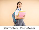 Small photo of Happy Indian student modern schoolgirl wearing school uniform holding books and bag standing isolated over beige background, Studio shot, Education concept.