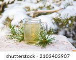 Hot pine tree needle tea infusion in transparent glass tea cup on table. Snowy pine tree on background, outdoors on cold winter day.