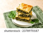Small photo of Stack of Spanakopita piece of pie, homemade Greek spinach pastry with cheese feta, chopped spinach, green onion, egg, layered in phyllo or filo dought. Light beige background with green napkin.