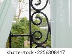 Small photo of Looking out a rain streaked glass window framed by gauzy sheer curtains to water drops on a wrought iron scrolled gate toward a background of rainy white sky, green, brown and peach in distance.
