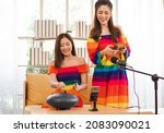 Two young women in rainbow color dresses enjoy traditional African music instruments kalimba and steel tongue drum vlogging video blogger. Beautiful girls have fun relaxing at home singing and playing