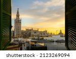 Small photo of View through an open window with shutters of the Great Cathedral and Giralda Tower of the Andalusian city Seville, Spain under a cloudy colorful sunset.