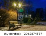 Old wooden bench dusted with first snow is illuminated by street lamp. City park in evening