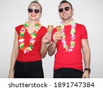young woman and man posing near white wall with glass of champagne in hand. boy and girl wearing hawaiian flowers and have PF21 written on foreheads. celebrating new year 2021 at home quarantine.