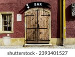 Small photo of stipple textured exterior stucco wall elevation closeup with old wood double wing gate. wood window and stone trim. fake or sample white BAR banner sign above on metal plate. classic architecture.