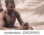 Small photo of A moment of leisure and tranquility as a young man sits under the shade of a palm tree, checking his phone with a nonchalant expression. The warmth and beauty of the tropical environment