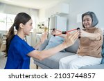 Small photo of Asian mature older woman doing physiotherapist with support from nurse. Senior elderly female patient sitting on sofa in living room use resistance stretch band exercise with caregiver in nursing care