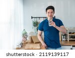 Small photo of Portrait of Asian attractive male holding a glass of water in kitchen. Young thirsty handsome man pouring and drink clean mineral natural in cup after wake up in early morning for health care in house
