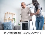 Small photo of Asian young daughter support senior older man walk with walker at home. Beautiful girl help and take care of elderly mature grandpa patient doing physical therapy for health in living room in house.