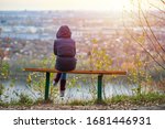 Small photo of Young woman sitting on bench in autumn city park and looking at cityscape, back view. Self-reflection of girl, thinking about unrequited love for Valentines Day. Introspection concept.